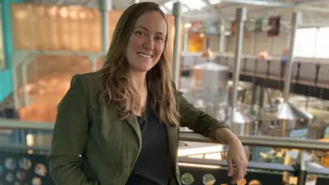 Alex Pope/BBC The image shows a person standing indoors inside a brewery on an upper-level walkway or balcony, resting one arm on the railing. She is wearing a dark green blazer over a black top. Her hair is long and light brown, falling over her shoulders. There are large metallic tanks, pipes, and equipment behind her, with high ceilings and plenty of natural light coming through large windows.