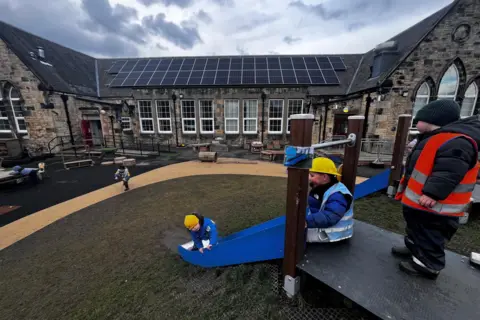 Young children play on a slide in the playground with the school building behind them which has solar panels on the roof.