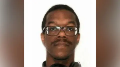 US Department of Justice Brian Jones Jr looking straight ahead and wearing square framed dark-rimmed glasses. He is against a plain white background. 