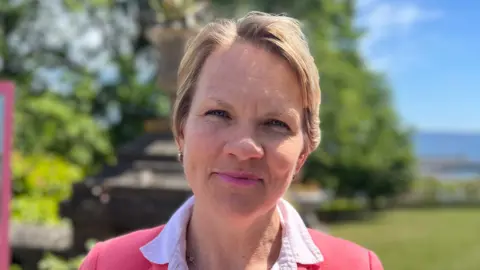 Sasha Kazantseva-Miller, a woman with short blonde air and a pink suit and shirt, smiles at the camera.