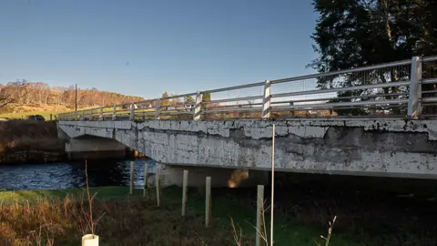 A crumbling road bridge in the Highlands on a sunny day with trees and fields in the background
