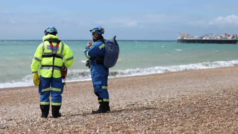 Two coastguard officers in yellow and blue on stoney beach with Brighton Pier in background