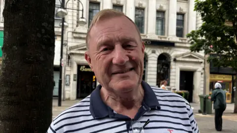 Gemma Dillon/BBC A man wearing a blue and white striped T-shirt is stood on a street. He has gingery brown hair and is smiling.