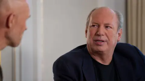 Hans Zimmer, wearing a blazer, sitting down speaking in an interview