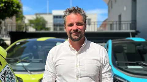 Nick Perchard-Rees is a man standing in the sunshine, smiling into the camera. He is standing in front of a green and a blue EVie hire car.