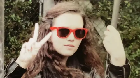 Family handout A photo of Maeve doing a peace sign with her right hand and holding onto a swing with her left, she is wearing red sunglasses and has brown curly hair