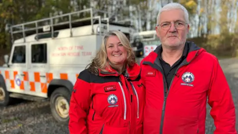 BBC volunteers Rachel Smith and Mark Silmon, wearing red overalls in mountain rescue uniform, stand outside a 4x4 rescue vehicle. They both smile looking into the room. 