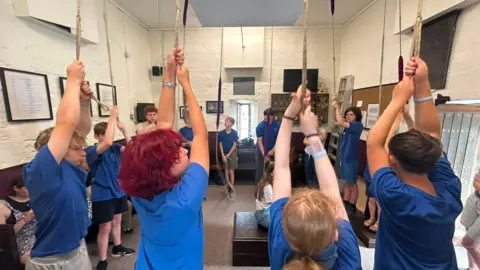 A group of young people are standing in a circle in a bell ringing tower. They are holding onto the ropes which are hanging from the ceiling above their heads. Most of the group are wearing blue T-shirts.