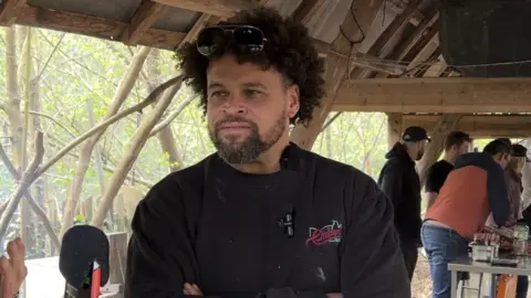 BBC A man with dark hair and a beard is wearing a black top. His sunglasses are on his hair and people are in the background.
