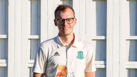 Contributed Luke Terry stands in front of a cricket club pavilion. He wears cricket whites and glasses. He has short, light brown hair.