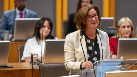 Eluned Morgan wearing a white jacket, addressing the Senedd, with Labour members sitting behind her