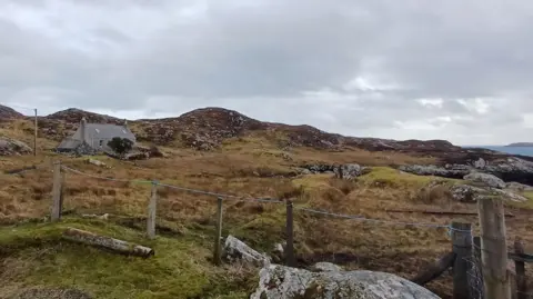 The small, single-storey house sits in a landscape of moor and bare rocks. There is a wire fence in the foreground.