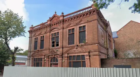 A two-storey building behind a fence. Trees and another structure are on the far left of the image.