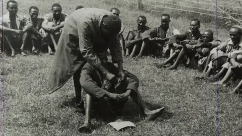 Bridgeman images Government de-oathing ceremony, 1953 - A Mau Mau detainee sitting in the centre of a circle of men in a fenced compound is purified of the Mau Mau oath.