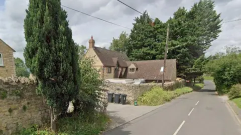 Google Part of Oxford Road in Garsington, showing a tree-lined road with detached properties surrounded by traditional brick walls