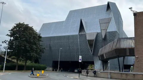 A large grey building known locally as The Elephant goes above a road in Coventry city centre. The side of the building most visible on the photo includes two large sections that are at different angles to each other.