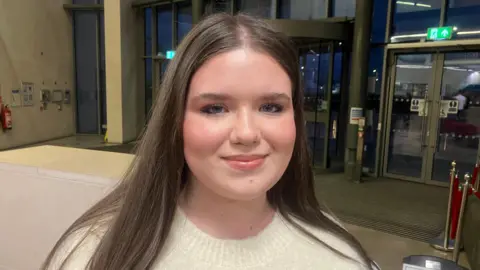 BBC A girl with long brown hair and blue eyes, wearing a white jumper. Behind her is a set of doors with windows.