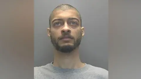 Cambridgeshire Police A police custody photo of Liam Rush, with short brown hair and a beard. He is wearing a grey T-shirt and is standing in front of a grey wall. He is looking above the camera.