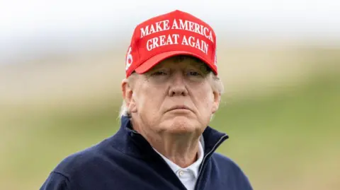 Head and shoulders of Donald Trump, who is wearing a white polo shirt, a navy blue fleece and a red cap which says "Make America great again"