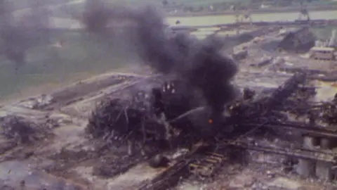 Burnt out chemical plant with black plume of smoke emitting from a burning fire at the centre