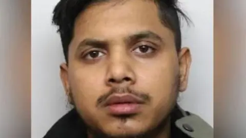A police mugshot of Jamil Talukder. He has brown eyes, a dark moustache and thin dark hair on his chin and cheeks.