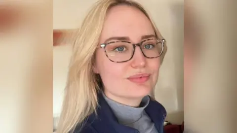 Name for Oxevision cameras to be banned in psychological well being sufferers' bedrooms Sophina A woman with long blonde hair wearing glasses is looking to the side of a camera. She is wearing a blue top over a grey roll neck top.