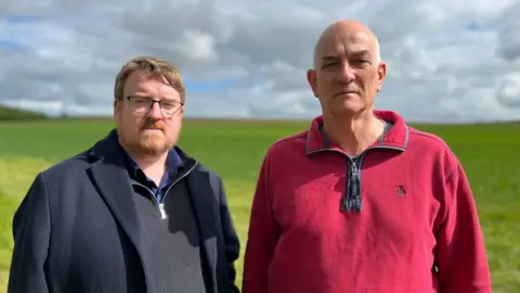 BBC Joe Corrall and John Greenhalgh from Hinchingbrooke Residents' Association standing next to each other with a field behind them. Joe has medium-length brown hair, glasses and a beard, and is wearing a navy coat and fleece. John is wearing a red quarter-zip jumper.