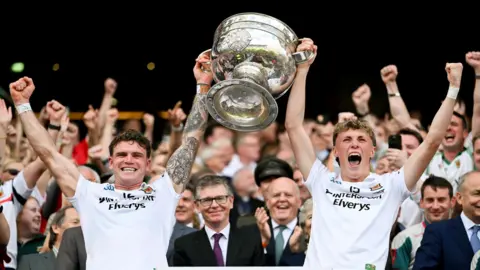 Sportsfile via Getty Images Two young men are standing with their backs to a large crowd in a stadium. They are both wearing white football tops and are holding the large, silver Sam Maguire trophy up between them. They are shouting and celebrating. 
