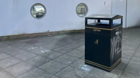 A black Newcastle CIty Council bin in the bust station. It has been graffitied with a tag in silver spray paint. Smashed bottles litter the floor around it.