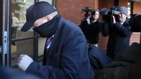 PA Media A man wearing a black baseball cap and black face covering and dark suite walks into a court building. He has a black hold-all on his shoulder.