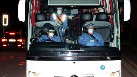 Getty Images The front of a coach carrying 20 Britons to Arrowe Park Hospital. Sitting at the front are three people wearing personal protective equipment including face shields, face masks, gloves, and coveralls. The tops of some passengers heads wearing face masks can be seen.