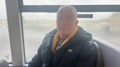 Maddy Jennings/BBC Ian Holman is sat on a grey leather seat, looking into the camera and wearing a navy coat with a yellow scarf. There is an emergency exit sign on the window behind him, and the window is dirty. To the left of the window there is a grey pillar.
