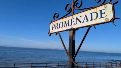 BBC The sign for the promenade in Maryport. It is weathered and mounted on a traditional wrought iron structure and has black writing on a white background. A clam blue sea sits behind it with dark peaks on the horizon.