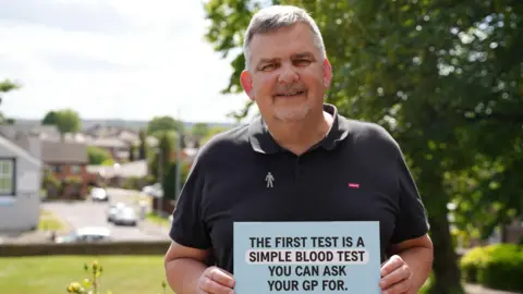 PCUK A man with grey hair and a beard stands in a garden. He wears a black polo shirt and a Prostate Cancer UK badge. He holds a card which reads: "The first test is a simple blood test you can ask your GP for."