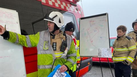 A firefighter holds a blue clipboard and points to a flipchart whilst other officers look on.