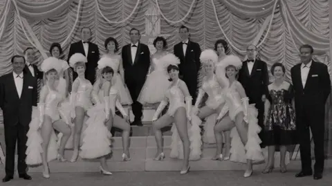 Manx National Heritage A total of 18 men and women stand on the stage in front of a ruffled curtain. Taken in black and white, the men wear black suits and white shirts and the women wear frilly white dresses, some have head dresses on.