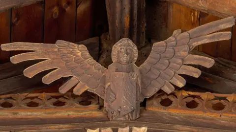 Mike Daines A wooden angel on the roof of St Wendreda's, March. Its wings our outstretched and it holding a shield in its hands. It is attached to a carved beam and above it can be seen the apex beam of the roof 