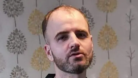 Adam Lawson is balding and has a beard. He is wearing a black T-shirt and is looking at the camera. He is standing in front of wallpaper with grey and gold trees on it.