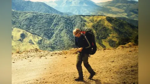 Karl Bushby A man with long blond hair walks along a rough mountain path. In the background are mountain peaks covered in trees.
