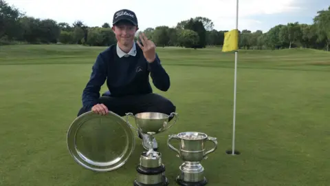 Family Handout A young boy wearing a navy blue cap, white polo shirt, jumper and trousers. He is crouched down on a golf course next to a hole which has a flag pole with a yellow flag in it. In front of the boy are two silver trophies and a platter. He holds the number three up with his fingers to signify the amount of awards he has won.