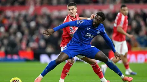 Getty Images Two footballers, one in a blue strip and one in a red strip, jostle over a yellow football on the pitch. 