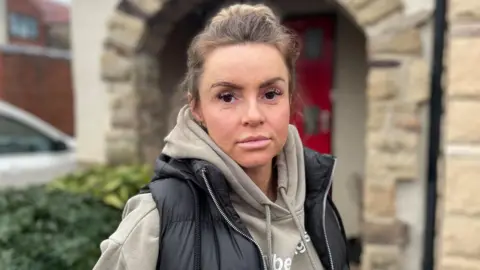 Simon Thake A young woman in a hoodie and a black gilet with her hair tied back. Her face shows a slight smile