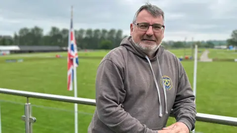 J&PR Ltd A man with short grey and brown hair and black glasses is wearing a grey hoodie with the blue and yellow Shrewsbury crest on its breast. He is leaning against a silver railing with glass panels. There is a large field with green grass below it, and a white flagpole with the Union Jack on it.