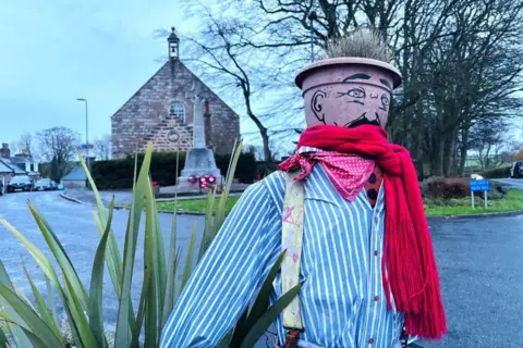A scarecrow-type character with a flowerpot head, red scarf and blue and white shirt and braces on a flowerbed, with a church in the distance.