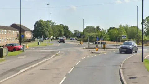 A road leading to a roundabout. Cars in the distance, trees on either side of the road. Houses on the left hand side of the road.