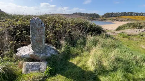 A stone memorial on the grass above a sandy beach, blue skies and calm waters in the background
