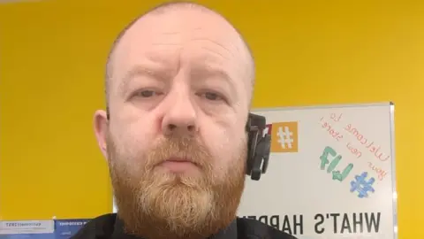 Geoff Savage Geoff Savage has a ginger beard. He is in front of a notice board and has an earphone with mic in one ear.