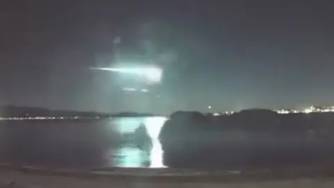 A green and white streak of light is reflected in water in a nighttime skyline.