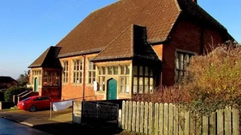 A photo showing the outside of a building. It has red bricks, a brown tiled roof and a green door. A red car is parked outside.