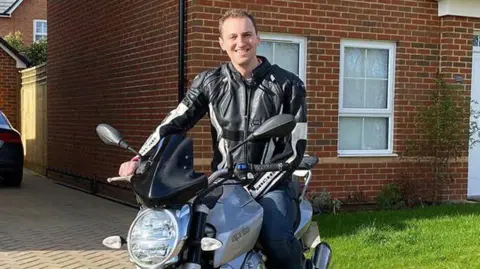 Peter Burch / Wiltshire and Bath Air Ambulance Charity A man with short hair is pictured smiling at the camera while sat on a stationary motorbike on a driveway. His right hand is on the handle of the bike. He is wearing a black leather jacket. 